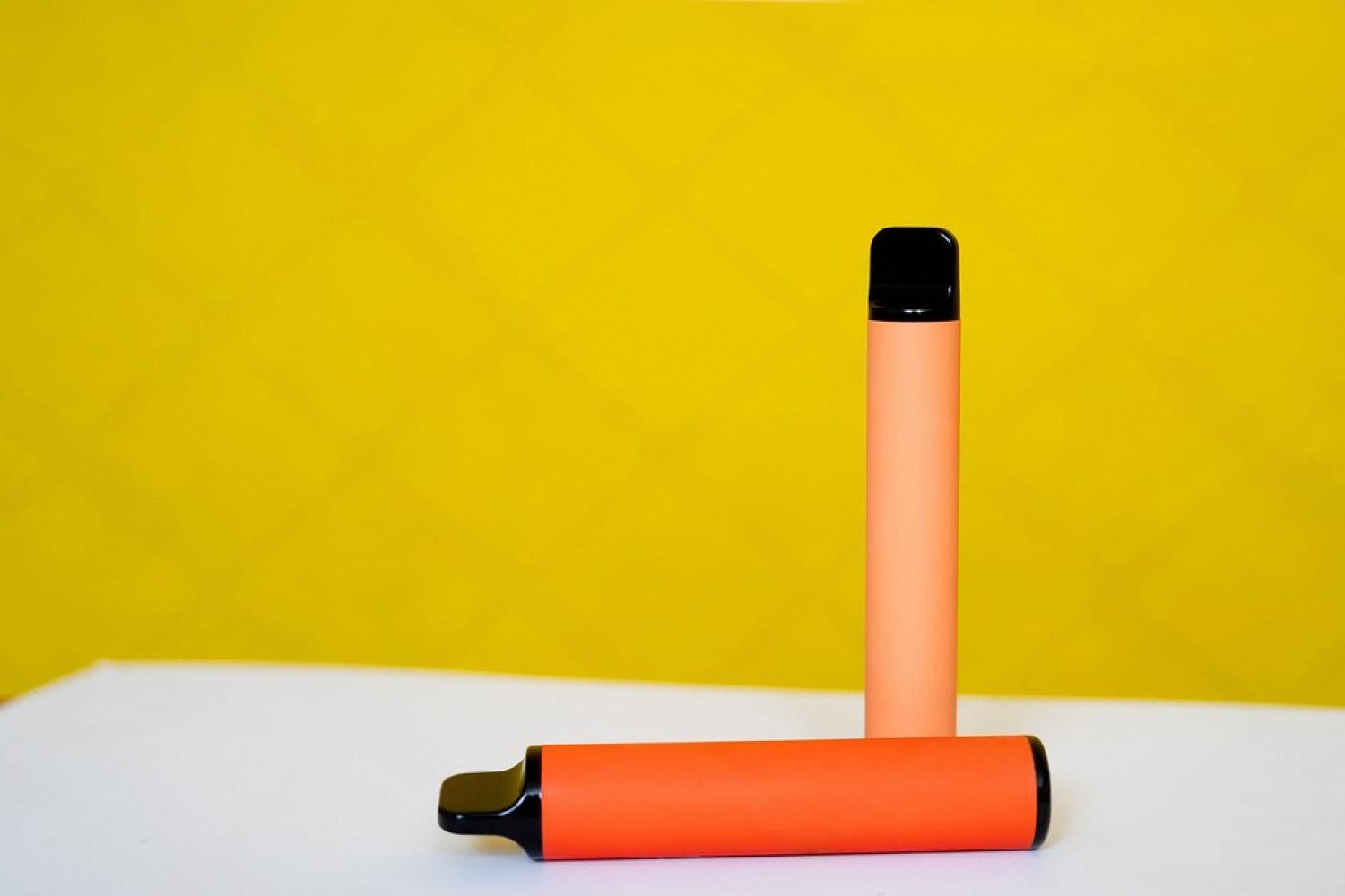 Two orange disposable vape devices standing on a white surface against a yellow background